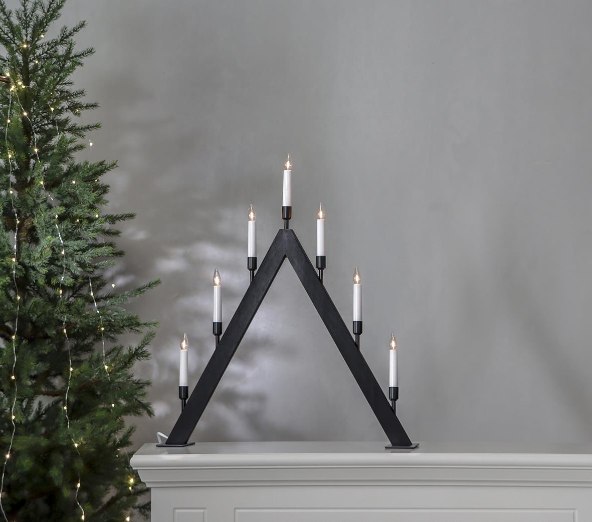 Svart ljusstake i triangelform med sju ljus, 55,5x63cm, placerad på en vit möbel bredvid en julgran