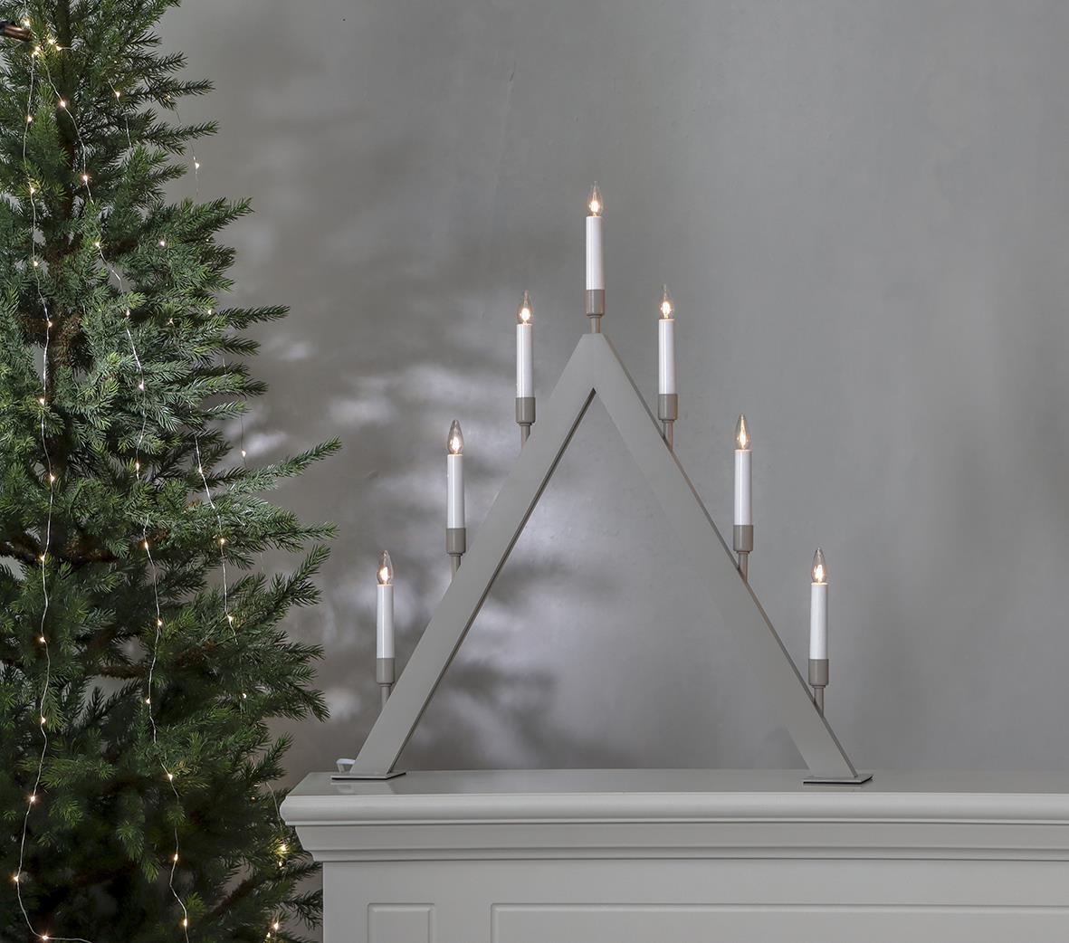 Ljusstake Vind Beige med sju ljus, formad som ett triangulärt träd, 55,5x63cm, placerad på en vit möbel bredvid en julgran.