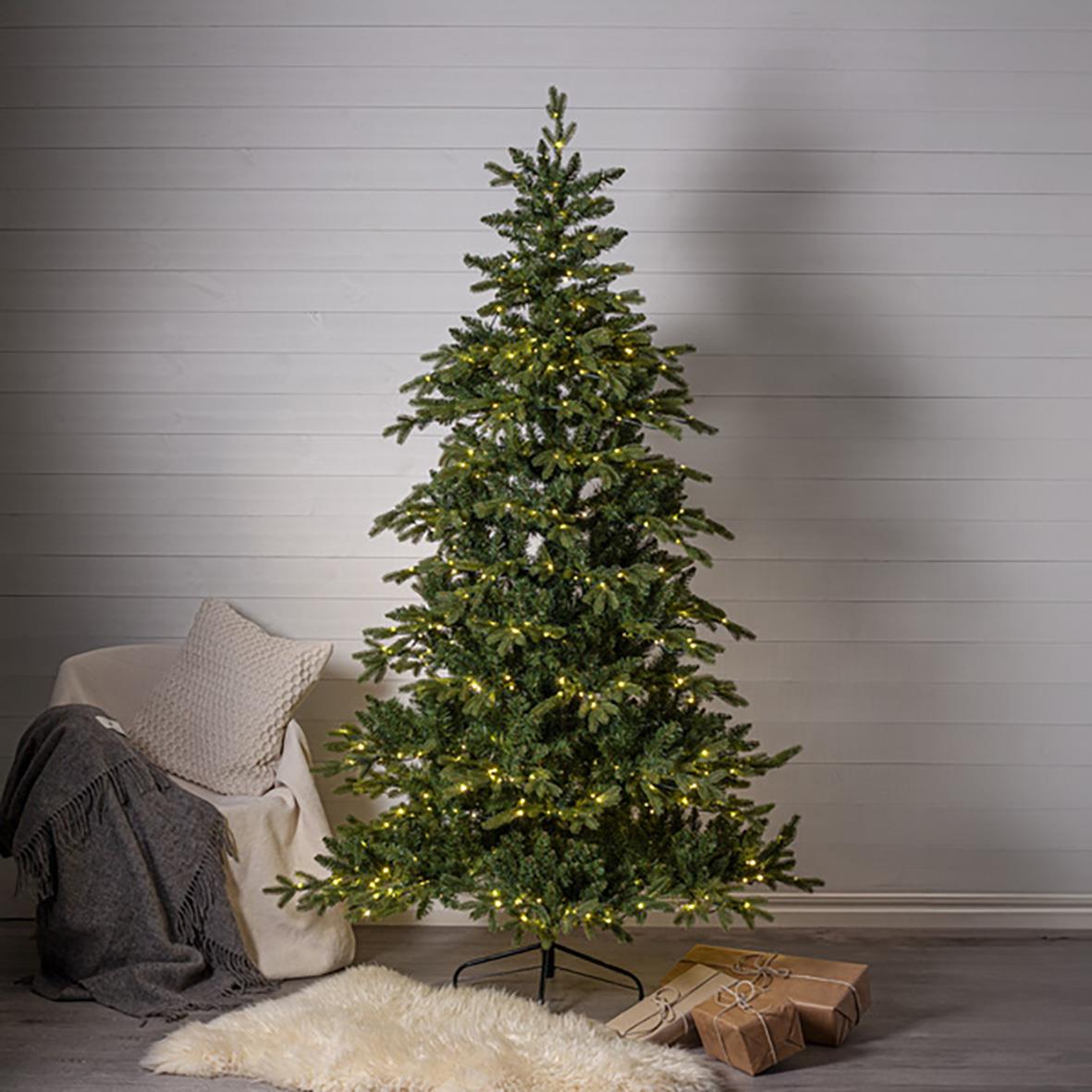 Julgran Larvik 700 LED lampor, 210 cm hög, står i ett rum med vit vägg, omgiven av julklappar och en filt på golvet