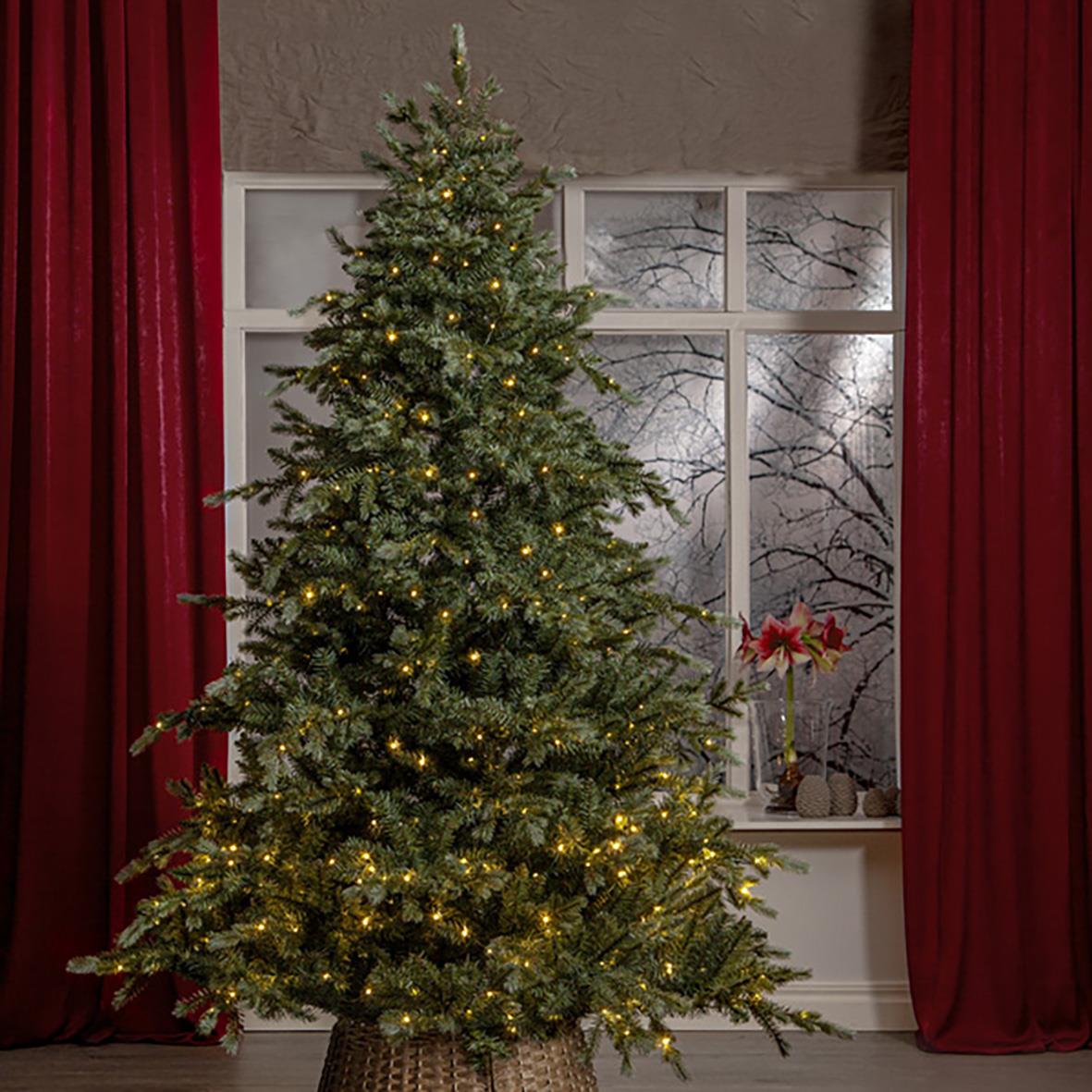 Julgran Greyland 700 LED-lampor, 210 mm, stående inomhus framför fönster med snölandskap utanför, omgiven av röda gardiner