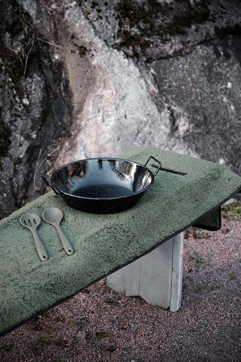 En svart stekpanna med vatten på en grön filt på ett bord utomhus, bredvid två plastskedar