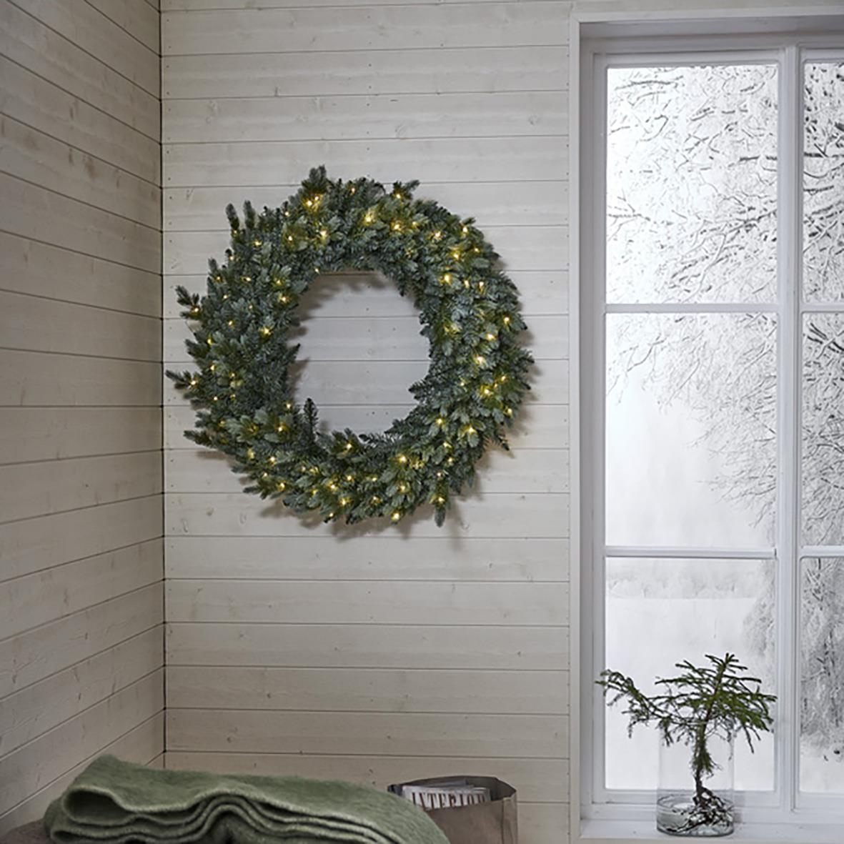 Grön krans med 80 LED-lampor, 100 cm, hängande på vit trävägg i ett rum med snö utanför fönstret