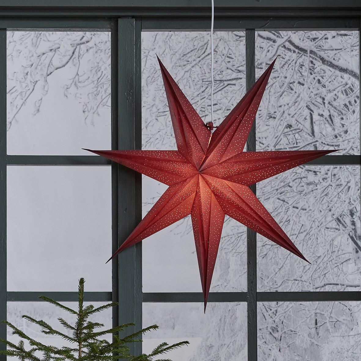 Röd julstjärna i papper hängande framför ett fönster med snöklädda träd utanför