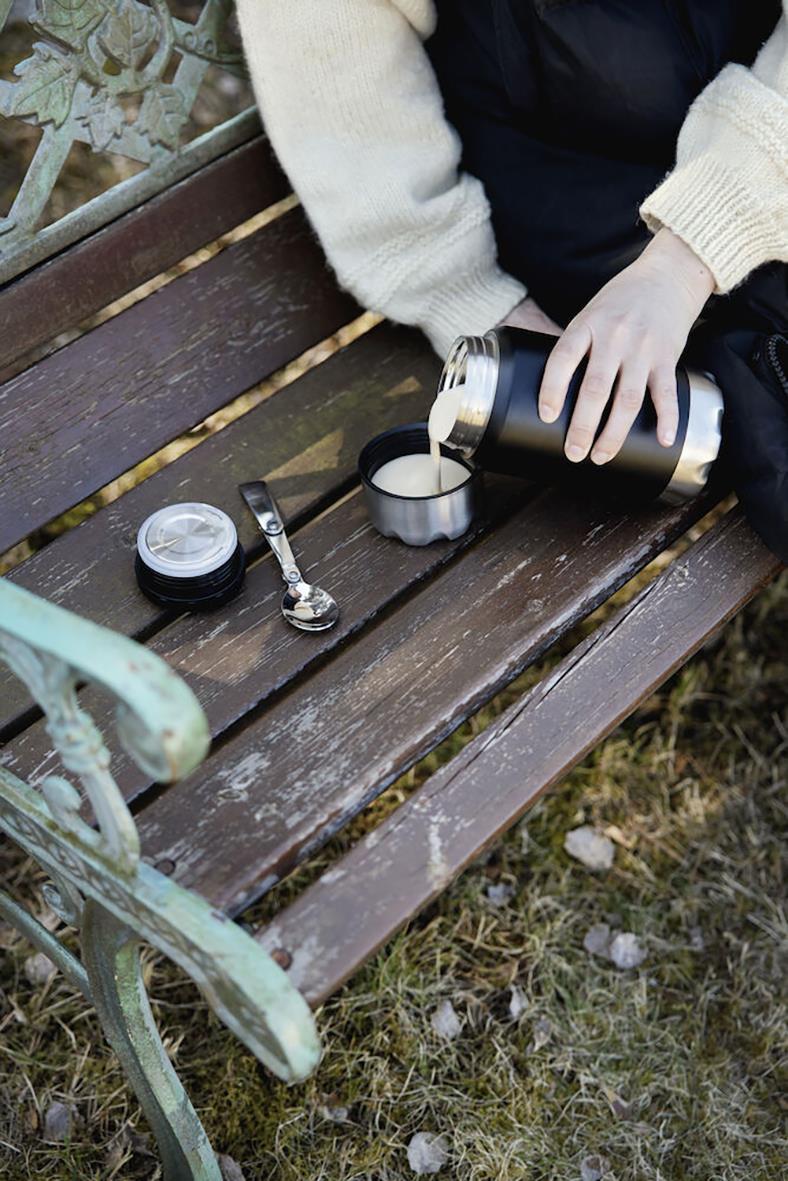 Person häller varm dryck från en svart termos i en kopp på en träbänk utomhus