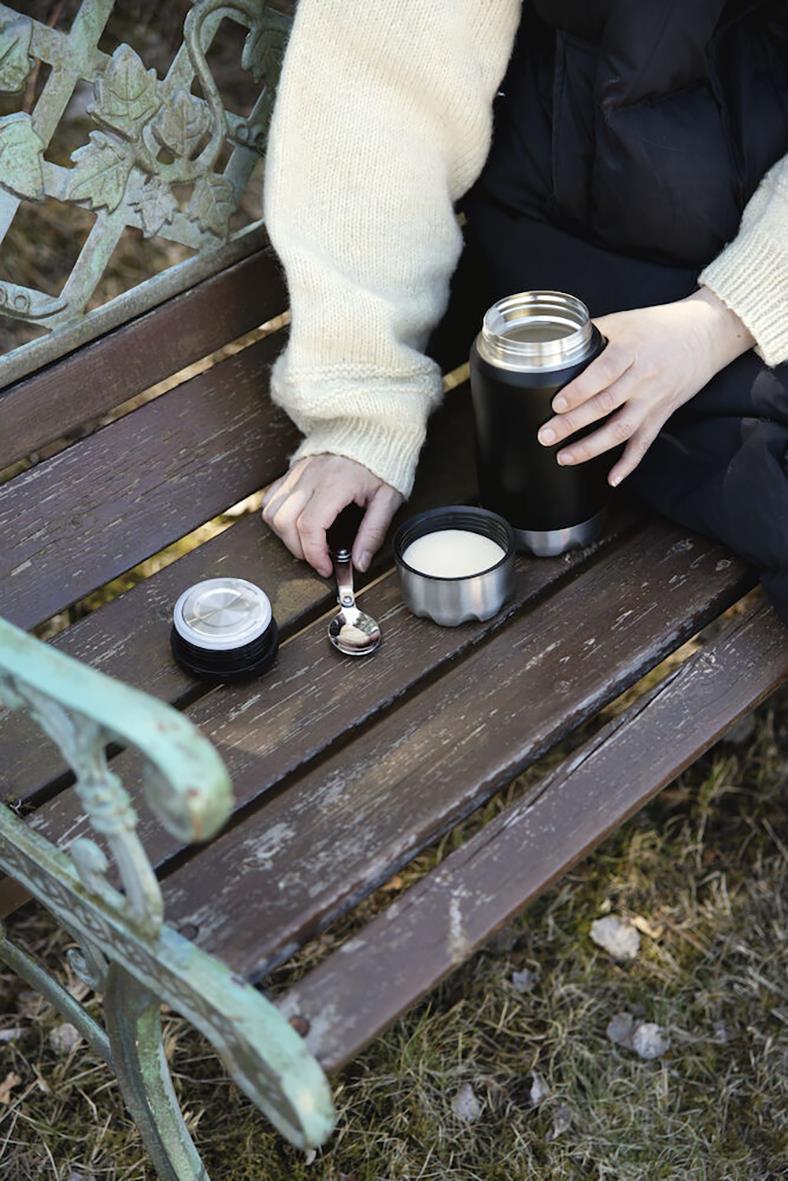 Person med vit tröja och mörk jacka använder en svart termos på en parkbänk, med ett lock och en sked bredvid.
