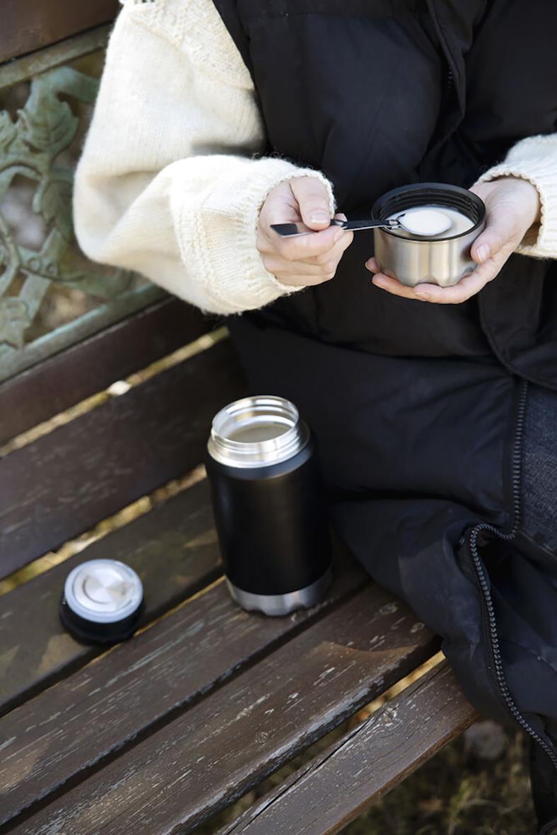 Person som håller en svart termosmugg med lock, sittande på en träbänk utomhus