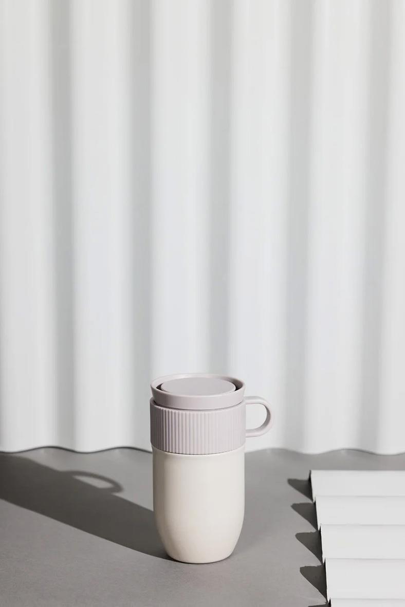 Beige termoskopp Sagaform Ted med lock och handtag, stående på bord framför vita gardiner