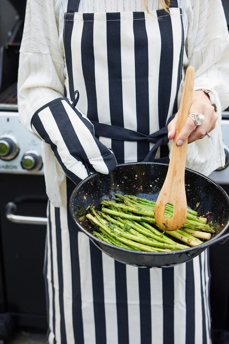 Person i svart/beige förkläde med grillvante lagar sparris i stekpanna utomhus