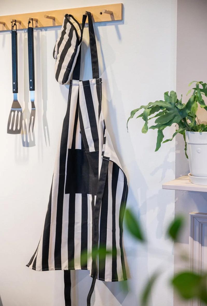 Förkläde med svart och beige randig design, hängande på krok i kök, med köksredskap och växt i bakgrunden