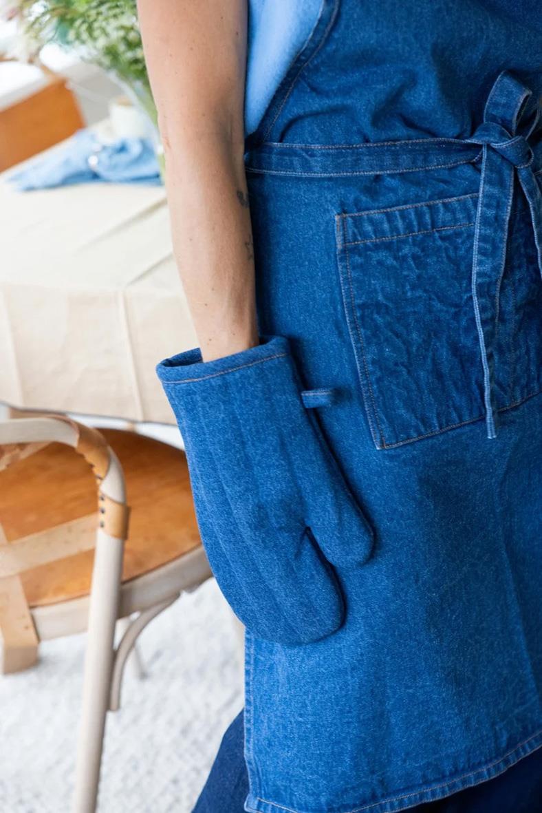Person bär en blå denimförkläde med ficka och knytband, sett bakifrån i ett hemmet.