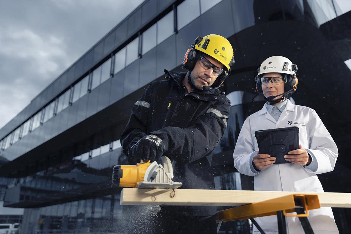 Två personer i skyddsutrustning med hörselkåpa och hjälm utomhus framför en modern byggnad