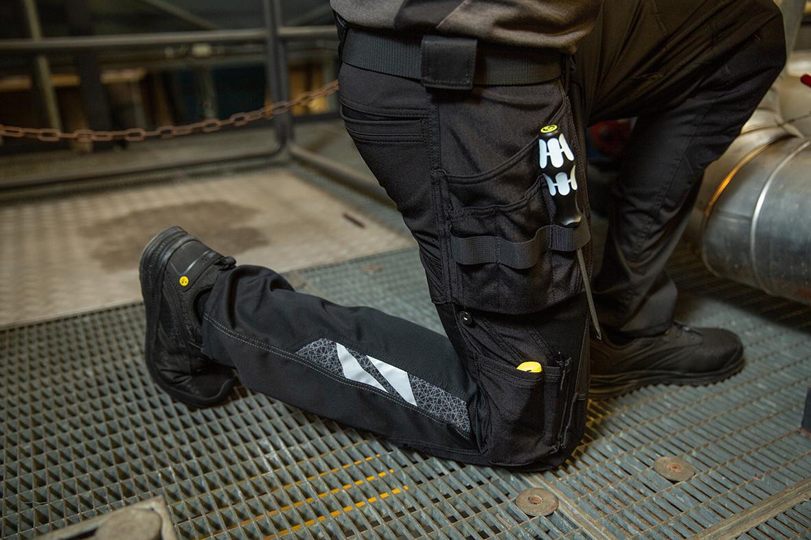 Person som bär svarta hantverksbyxor med verktyg i fickorna, knästående på en metallplattform i en verkstad