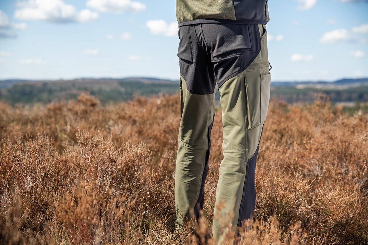 Person som bär mossgrön och svart unisex byxa i ett fält med buskar och himmel i bakgrunden