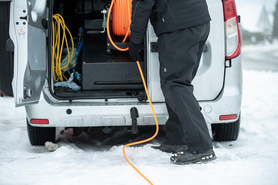 Person som kopplar en orange kabel till en svart arbetsbyxa framför en vit arbetsbil i snö