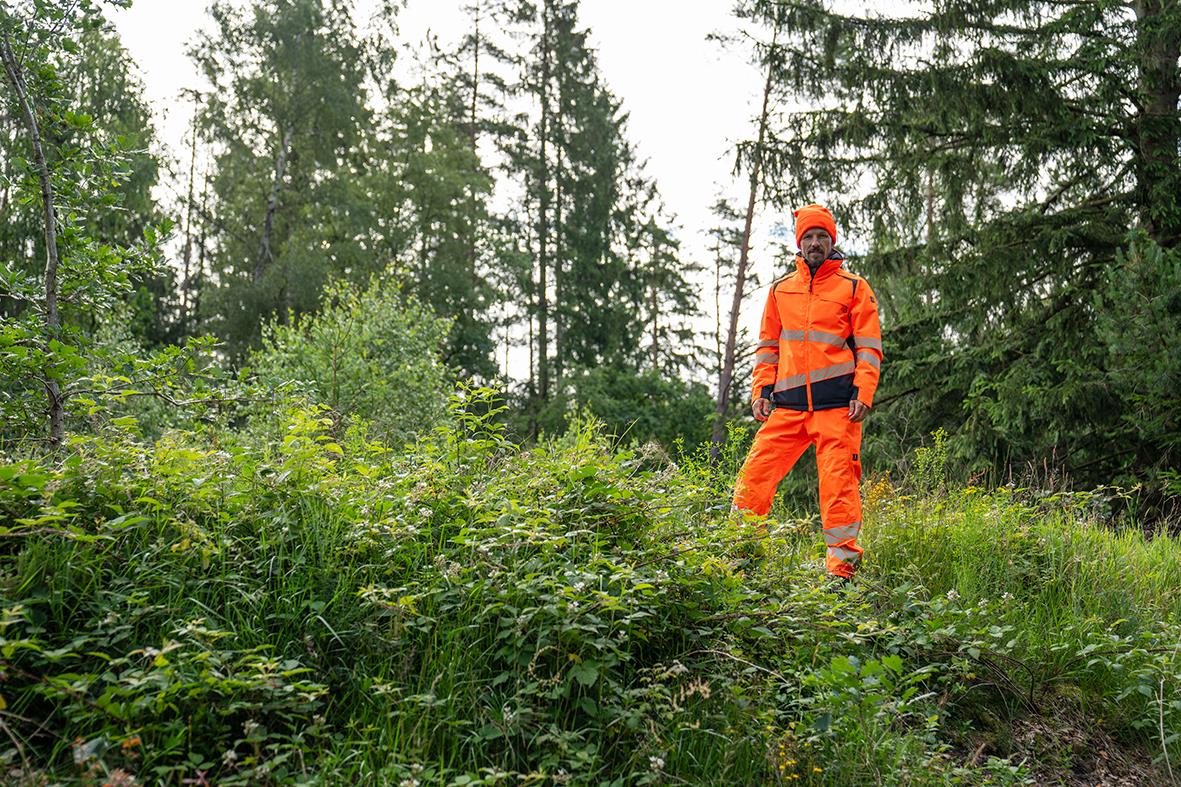 Person i orange överdragsbyxor och jacka står i skogsterräng med träd och buskar
