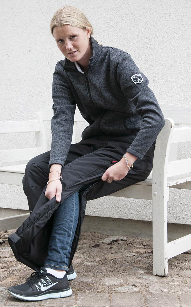 Kvinna sätter på sig svarta överdragsbyxor utomhus, sitter på en vit bänk, bär svart fleecejacka och sneakers