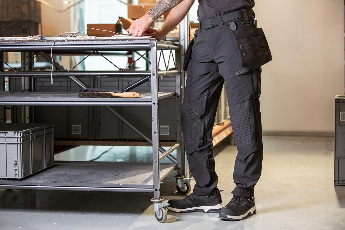 Person i svarta arbetsbyxor och sneakers står vid ett metallbord i ett arbetsrum