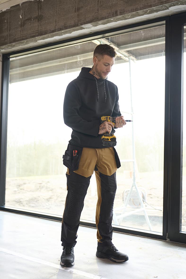 Man i svart hoodie och arbetsbyxor med verktyg, står inomhus vid stort fönster, använder skruvdragare