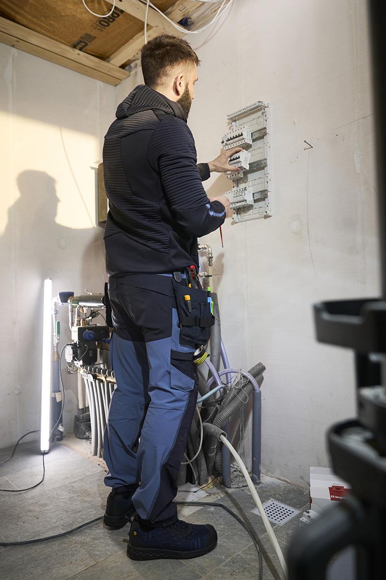 Man arbetar med elcentral i ett rum med byggmaterial och verktyg, klädd i arbetskläder och bärande verktyg i bälte