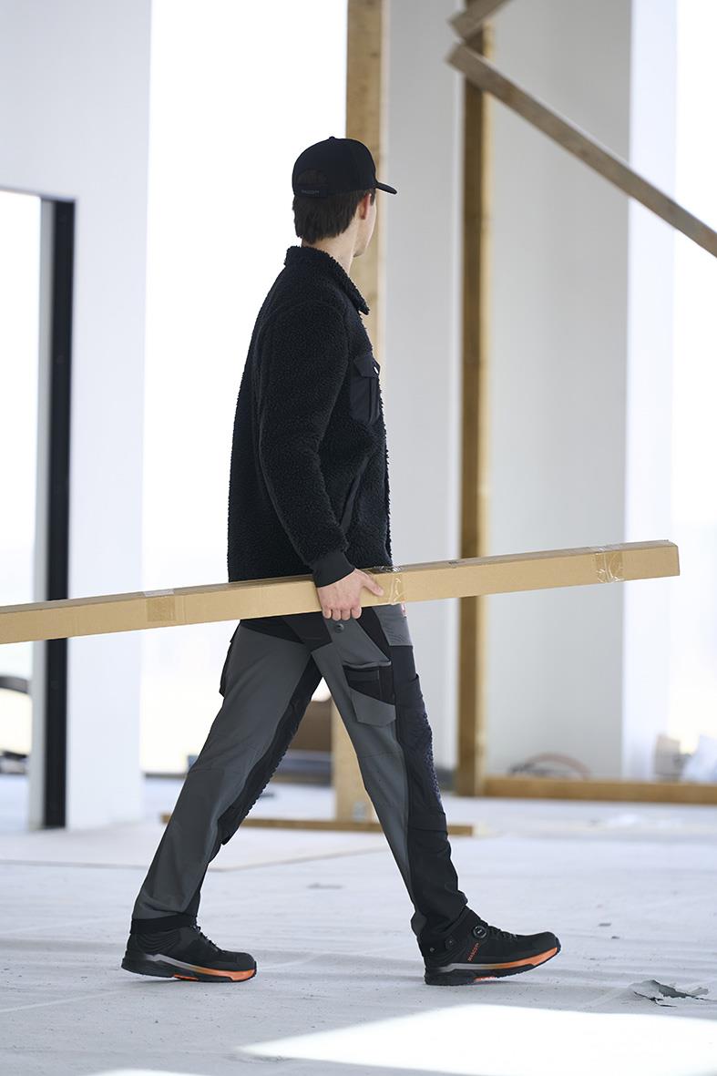 Person i svart jacka och svart keps bär en lång trästång i ett byggprojekt under konstruktion