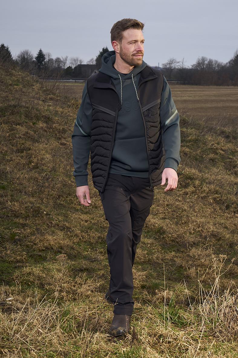 Man i svart funktionsbyxa och jacka går i ett öppet landskap med gräs och träd i bakgrunden