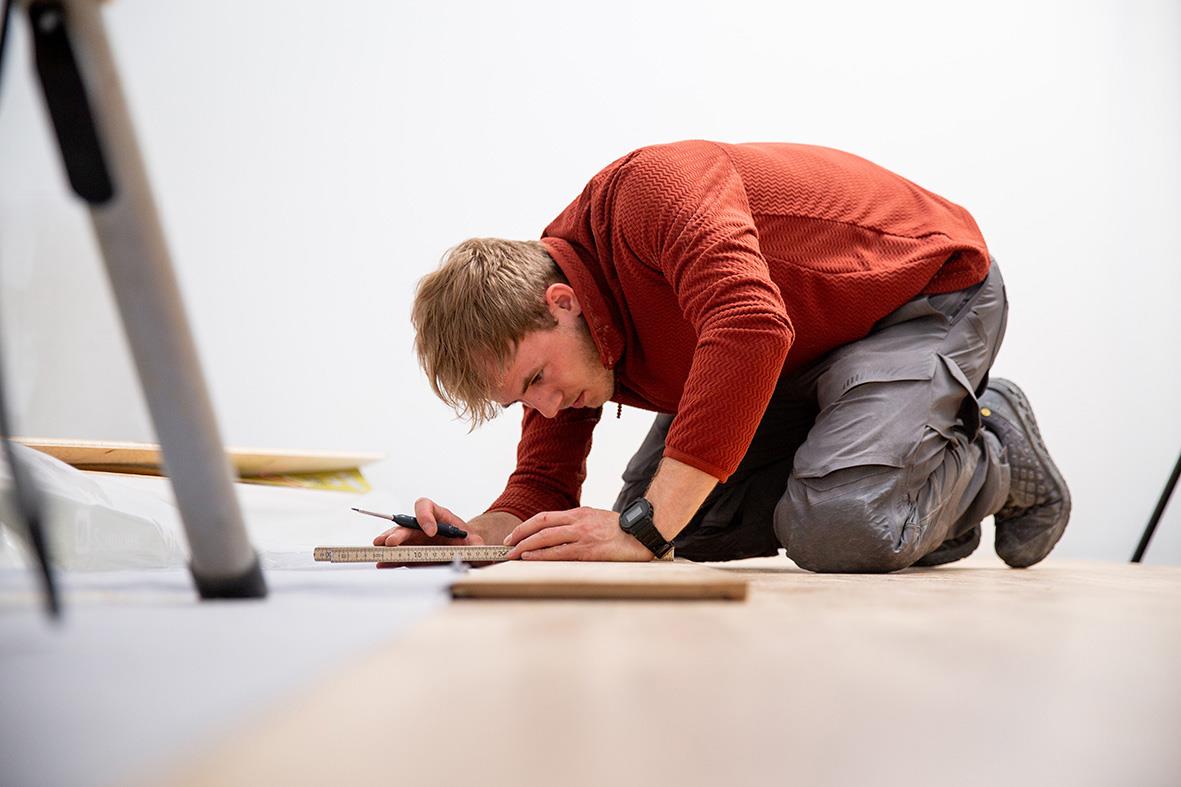 Man på knä på golvet, skriver eller ritar på ett papper, i ett rum med vit vägg och möbler