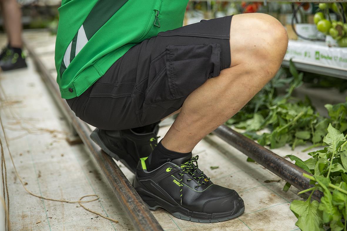 Person i svarta shorts och svarta och gröna arbetsstövlar, knäböjande i ett växthus