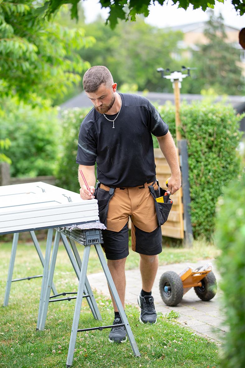 Man i svart t-shirt och bruna shorts står utomhus och skriver på ett bord, med en skateboard i bakgrunden