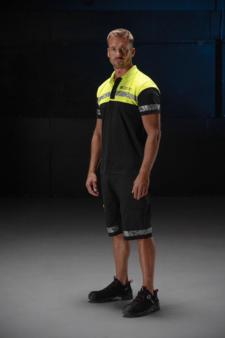 Man i svart och gul arbetskläder med shorts och tröja, stående på mörk bakgrund