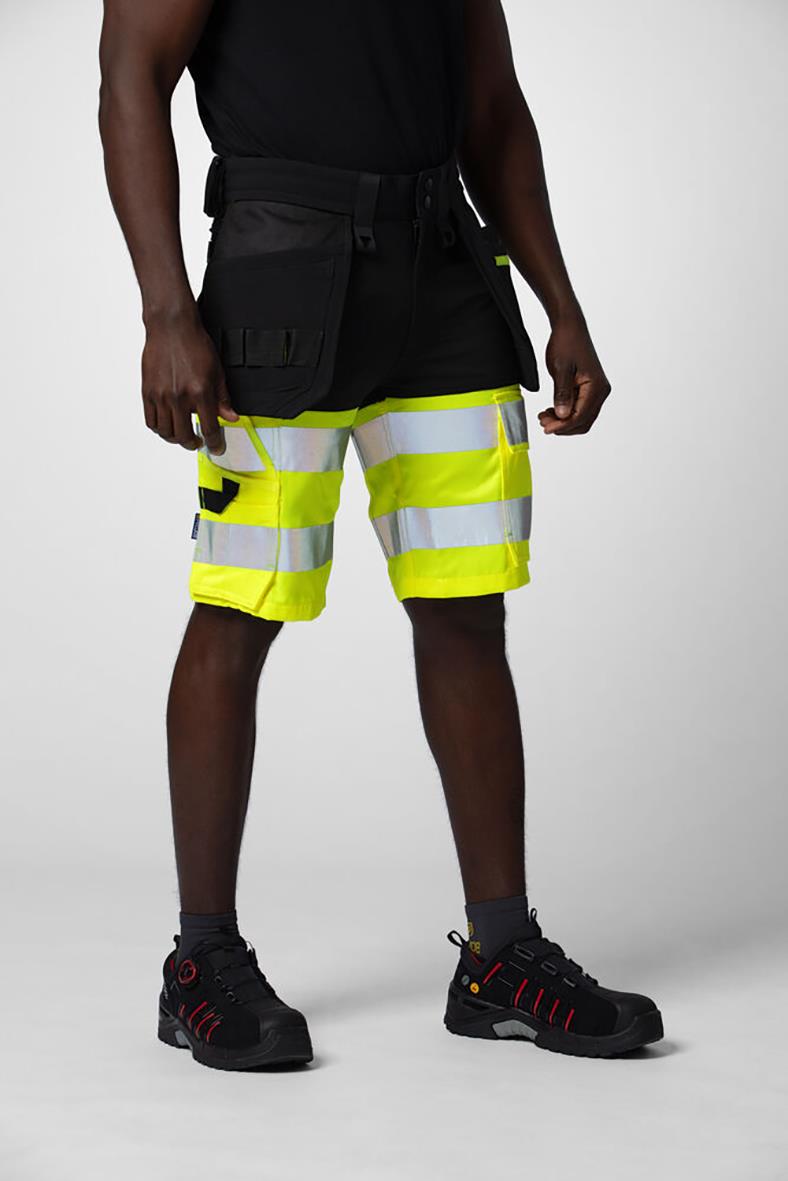 Person som bär svarta hantverksshorts med reflexer och verktygslåda, svart t-shirt, svarta arbetsstövlar, på neutral bakgrund