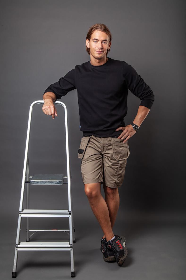 Man i svart tröja och khakifärgade hantverksshorts står bredvid en stege mot en grå bakgrund
