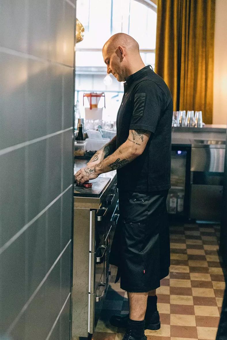 Man i svart t-shirt och shorts står vid köksbänk i ett kök med guld- och blå gardiner