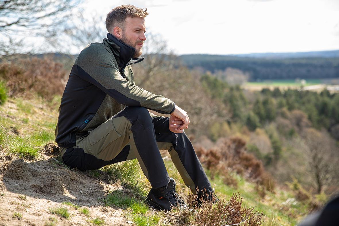 Man sitter på en klippa i naturen, bär en fleecetröja i mossgrön och svart, ser ut att njuta av utsikten