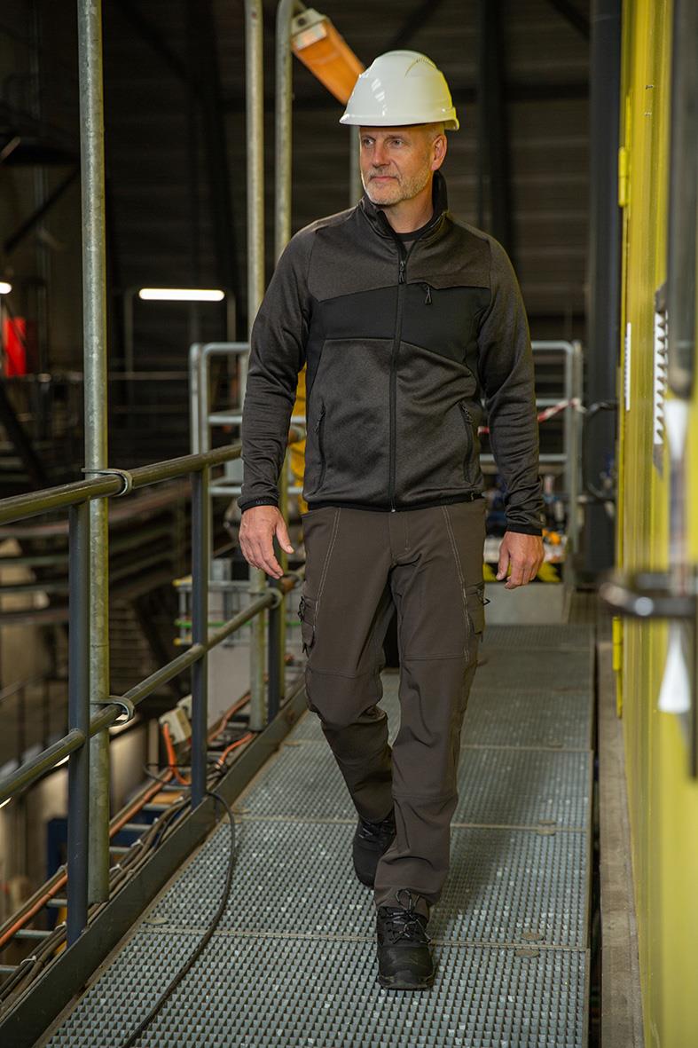 Man i svart fleecejacka och mörkgråa arbetsbyxor med hjälm i industrimiljö