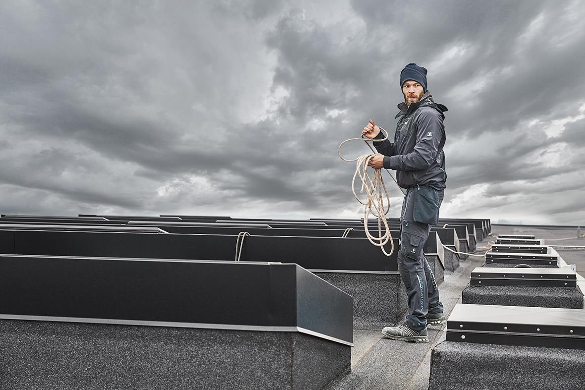 Man står på tak med mörk marin stretch skaljacka och mössa, håller i rep under en molnig himmel