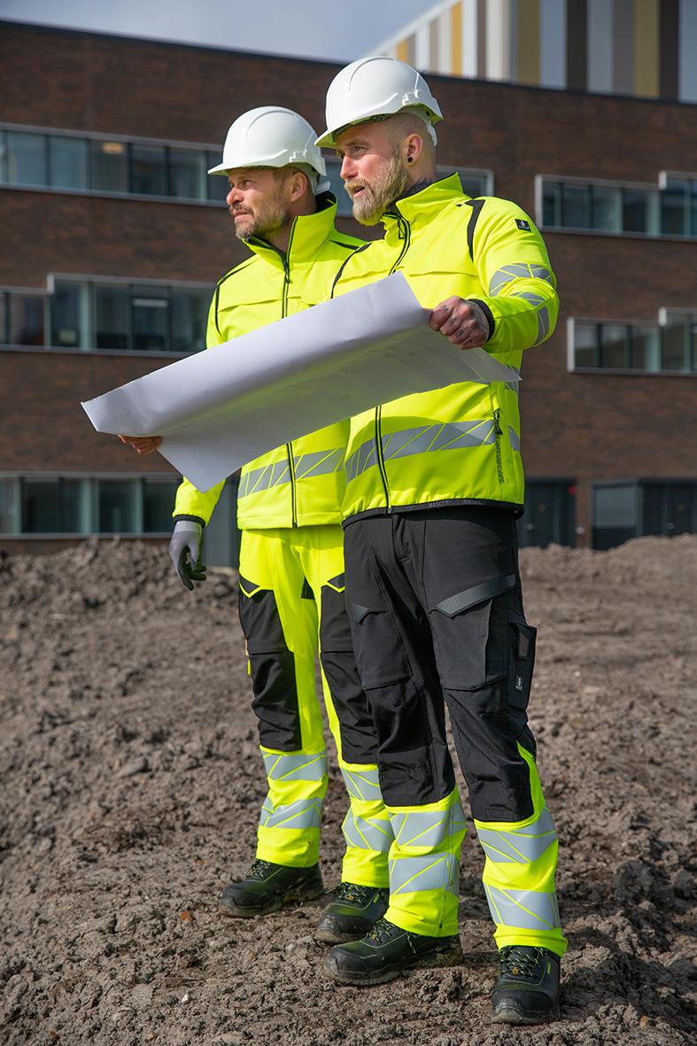 Två byggarbetare i varselgula jackor och vita hjälmar granskar en byggplan utomhus framför ett modernt kontorshus