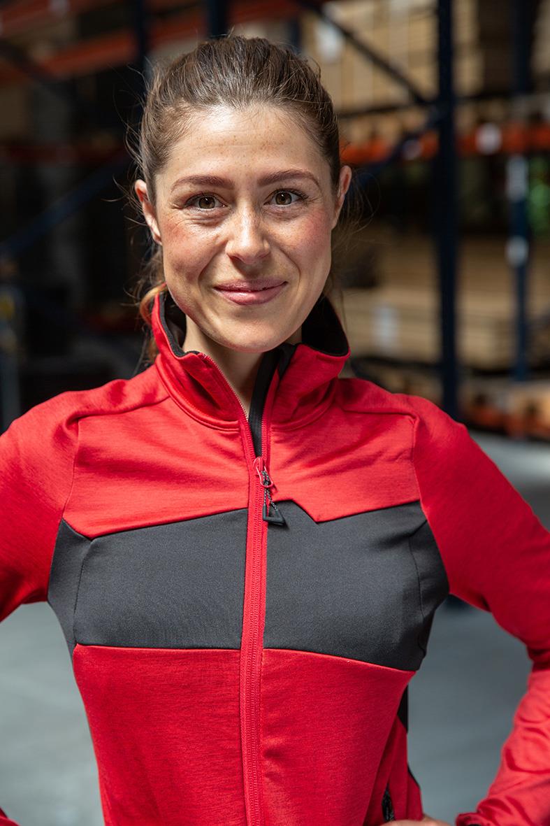 Kvinna i röd och svart fleecetröja med hög krage, stående i lagerlokal