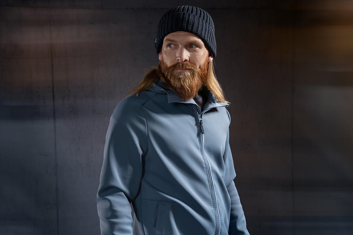 Man med långt hår och skägg i svart mössa och blå fleecetröja mot mörk bakgrund