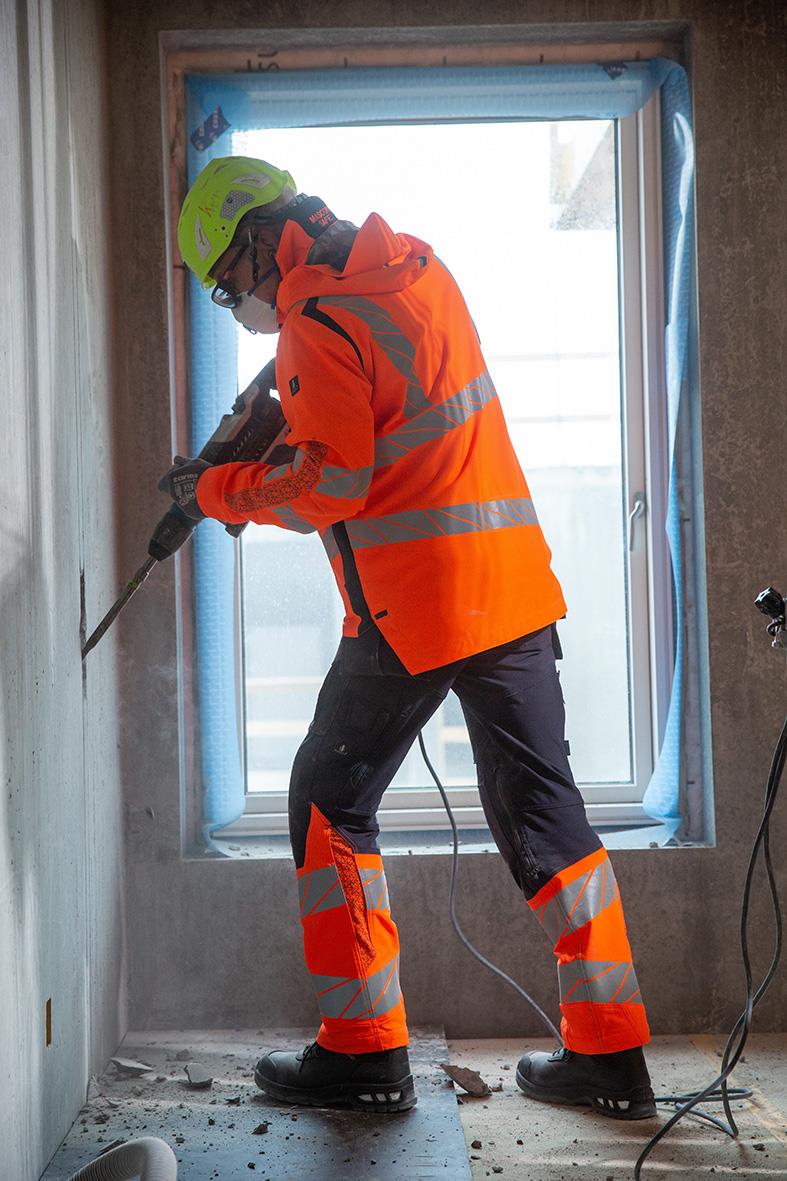 Person i orange varseljacka och hjälm borrar i vägg inomhus vid ett fönster