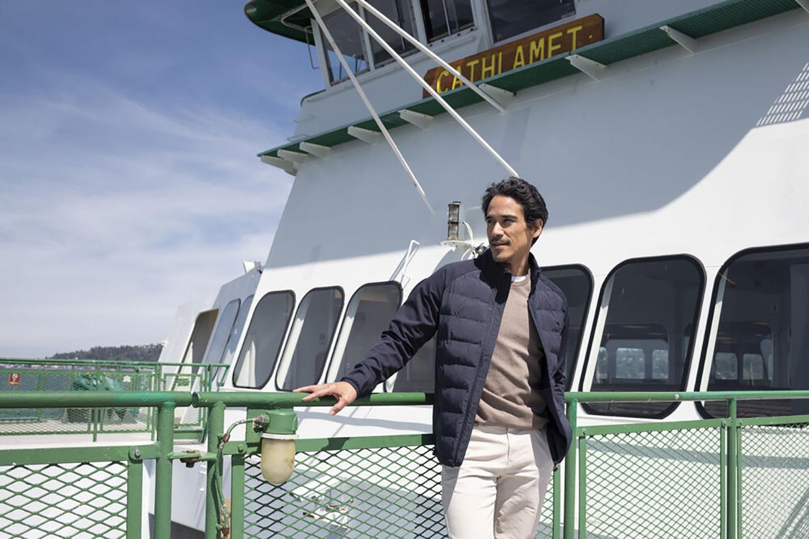 Man står på en båt med marinblå jacka och ljusa byxor, utsikt över vatten och himmel i bakgrunden