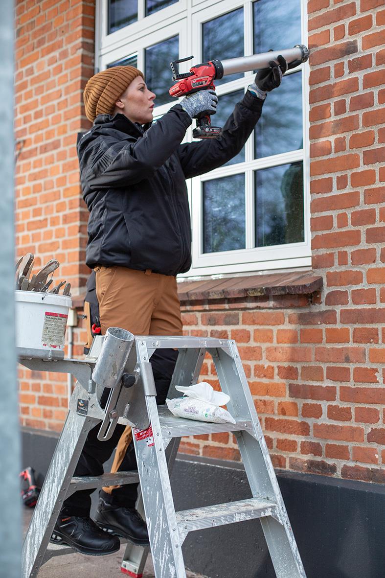 Kvinna i svart jacka och brunt mössa använder en borrmaskin på en stege utomhus vid ett tegelhus