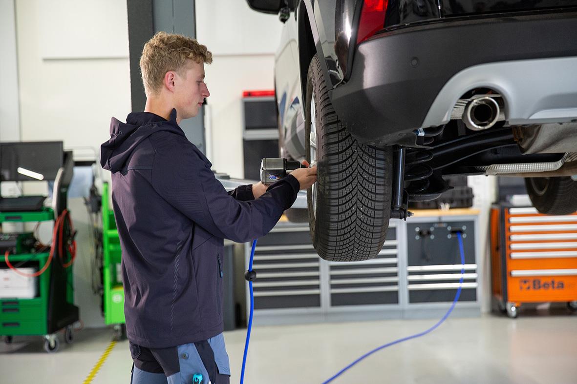 Ung man inspekterar ett fordon i en verkstad, bär en mörk marin jacka och använder ett verktyg.