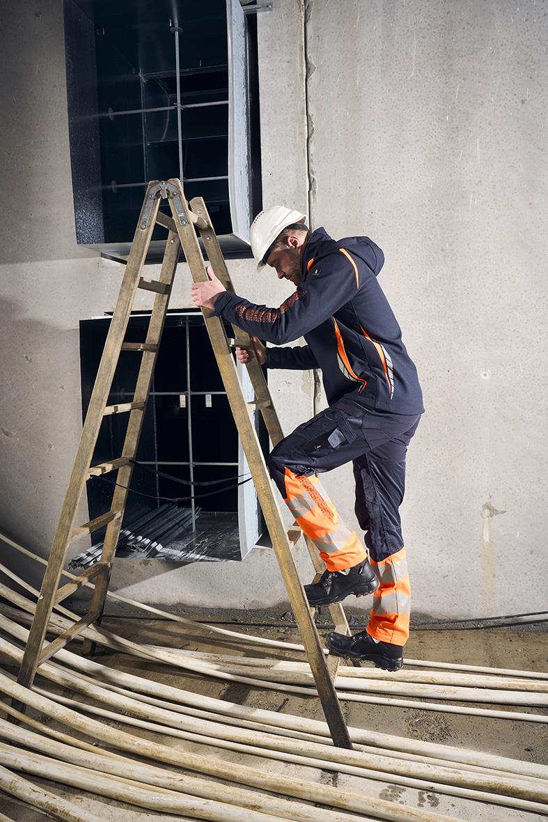 Person som arbetar på stege utomhus, bär mörk marin och orange arbetskläder, nära ett fönster och kabelrör