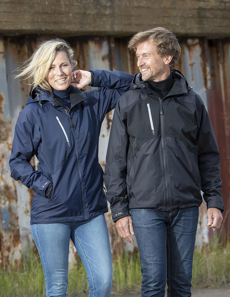 Två personer bär svarta och blå skaljackor utomhus framför en rostig metallvägg, båda ler och ser glada ut.