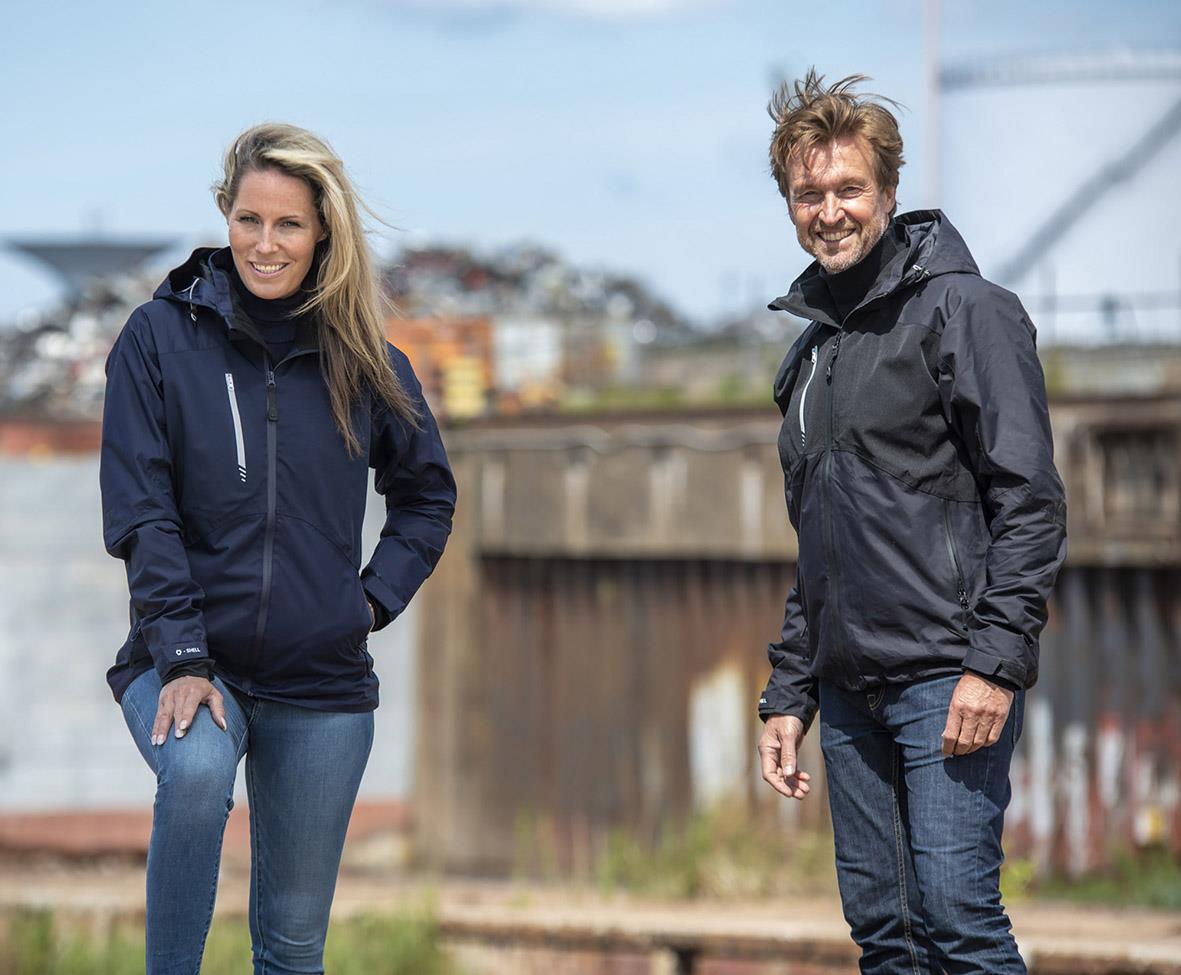 Två personer bär skaljackor utomhus, en kvinna och en man, med en stad och hamn i bakgrunden