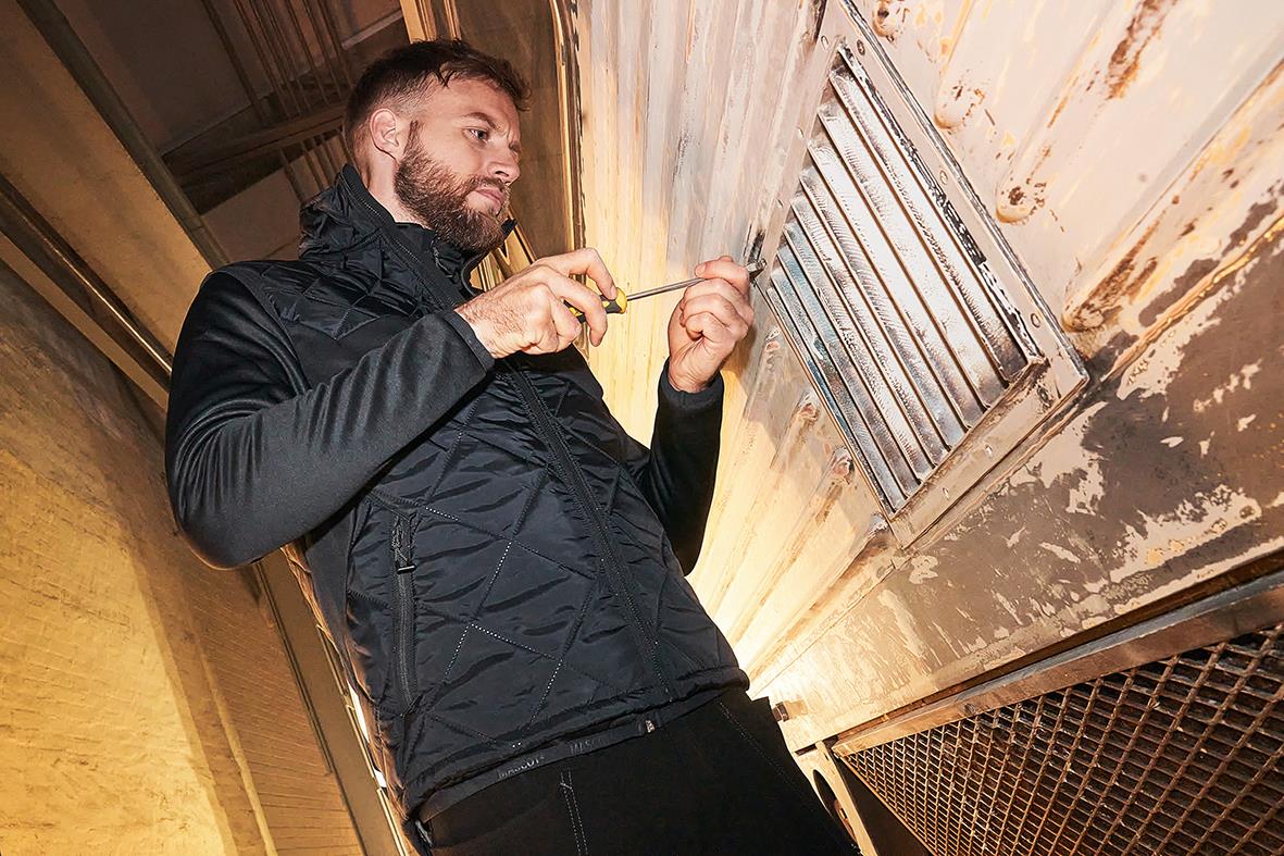 Man i svart vinterväst som tittar på en ventilationsgaller i ett äldre, slitet tak