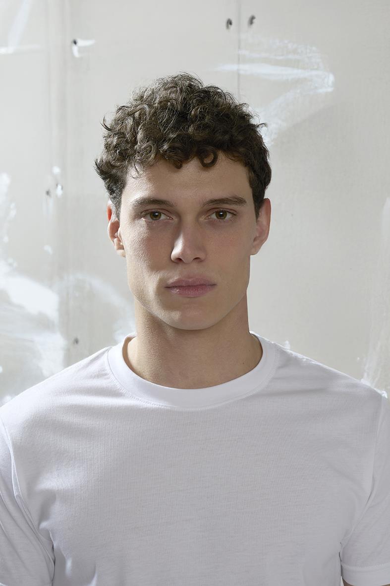 Ung man med lockigt hår i vit T-shirt står framför en ljus bakgrund