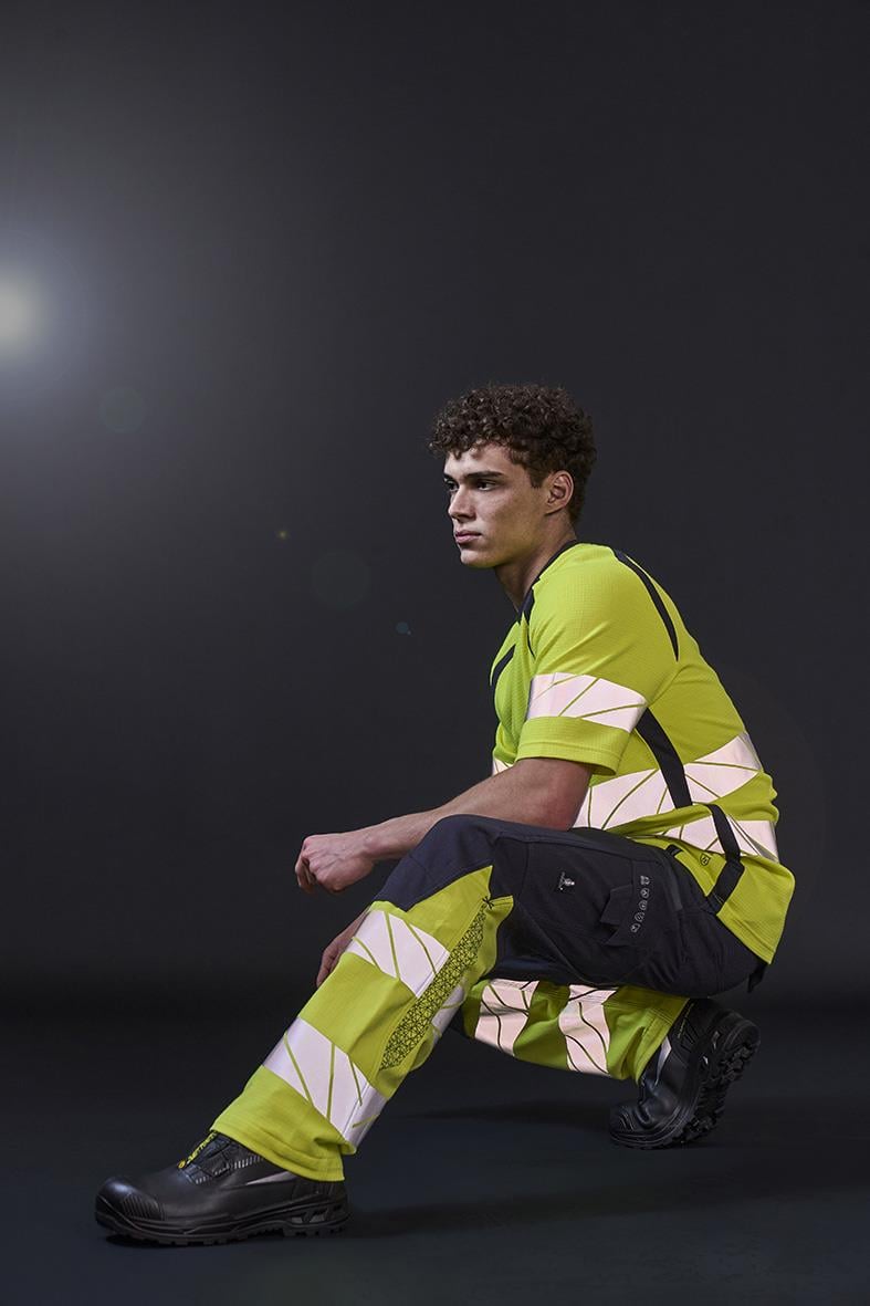 Ung man i varselgul och marin arbetskläder i knästående position mot mörk bakgrund