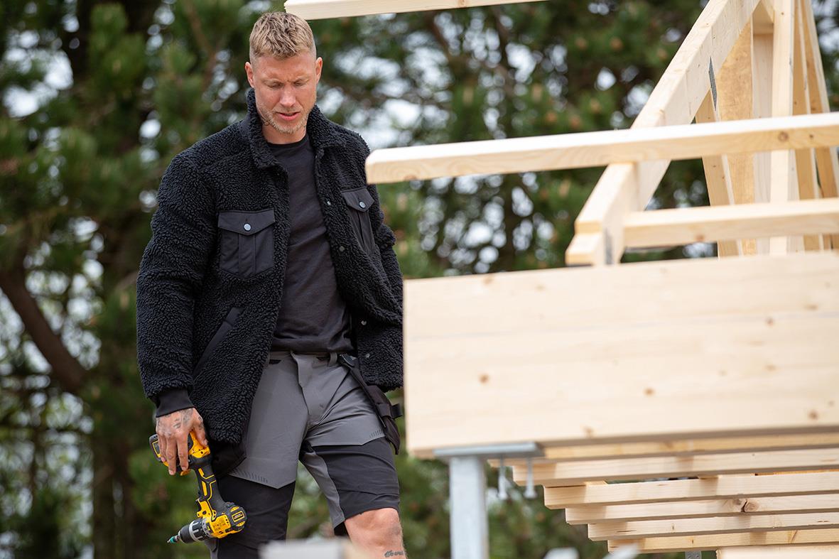 Man bygger ett trädäck utomhus, bär svart jacka och grå shorts, med verktyg i handen