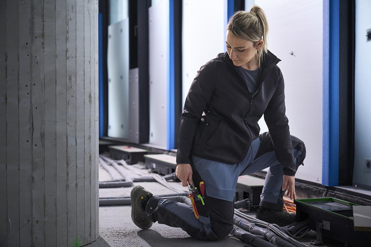 Kvinna i svart fleecejacka och mörk marinfärgade byxor arbetar med kablar i en industriell miljö